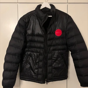 Svart pufferjacka från Hugo Boss - Svart pufferjacka från Hugo Boss med röd rund logotyp på bröstet. Jackan har quiltad design, hög krage och två sidofickor med dragkedja. Materialet är glansigt och syntetiskt, perfekt för kalla dagar. Stängs med dragkedja framtill. Finns även en avtagbar luva.