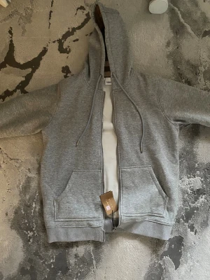 Grå hoodie från Burberry med rutig huva - Säljer en grå hoodie från Burberry i storlek M. Tröjan har dragkedja, snörning vid huvan och två fickor fram. Insidan av huvan har Burberrys klassiska rutiga mönster i beige, svart och vinrött. Perfekt för dig som gillar stilrena och lyxiga detaljer. Det står storlek M men egentligen är de för storlek L den e lite för lång!