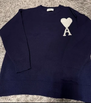 Marinblå stickad tröja från Ami - Säljer en marinblå stickad tröja från Ami med vit hjärtformad A-logga på bröstet. Tröjan har rund hals, ribbade muddar och långärmad passform. Först till kvarn!!!