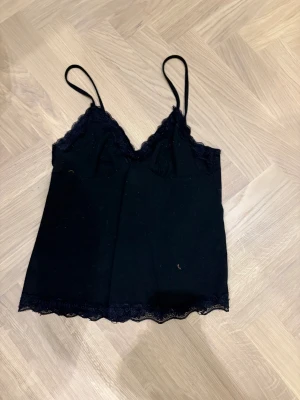 Svart spetslinnetopp från Romwe XS - Svart linnetopp från Romwe i storlek XS med tunna axelband och v-ringad halsringning. Toppen har fina spetsdetaljer längs urringning och nederkant, vilket ger en extra snygg touch. Perfekt att styla med jeans eller kjol för en trendig look.