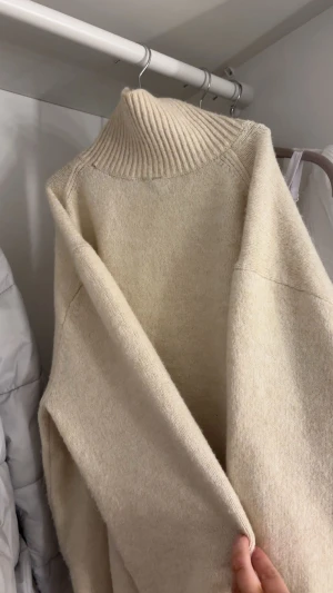 Beige stickad polotröja - Mysig beige stickad polotröja med ribbad hög krage och ribbade muddar. Tröjan har en loose passform och trekvartsärmar som kan vikas upp. Perfekt för dig som gillar enkel och clean stil med lite oversized känsla.