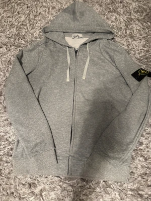 Grå hoodie från Stone Island M - Snygg grå hoodie från Stone Island med klassisk logopatch på vänster ärm. Tröjan har huva med snörning, dragkedja framtill och två fickor.