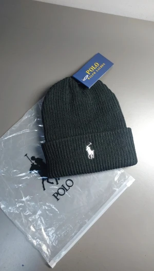 Svart mössa Polo Ralph Lauren - Svart ribbstickad mössa från Polo Ralph Lauren med klassisk broderad logga i vitt framtill. Mössan har uppvikt kant och är tillverkad i mjukt stickat material som håller dig varm och snygg. Perfekt accessoar för kyliga dagar.
