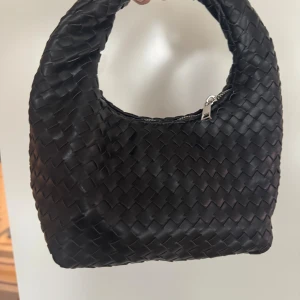 Gina Tricot flätad handväska, svart - Stilren flätad svart handväska  med rundad form och kort handtag. Väskan har dragkedja upptill och mäter ca 24x21 cm. Perfekt accessoar för dig som gillar en trendig och clean look. Det tvinnade handtaget ger en unik designelement till denna konstläderväska från Gina Tricot. 