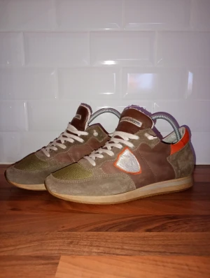 Philippe Model trpx - Snygga sneakers från Philippe Model i brunt, beige och olivgrönt med orange detaljer. Skorna har en rund tå, platt sula och snörning. Materialmix av mocka, skinn och textil. Ikonisk sköld på sidan i silver med orange kant. Perfekt för dig som gillar retrostil. Vid köp gratis marmor armband!