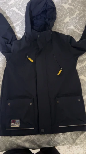 Svart vindjacka från Hampton Republic - Svart vindjacka från Hampton Republic med huva, två stora fickor med tryckknappar och två bröstfickor med gul dragkedja. Jackan har stilren design och detaljerad logotyp på ena fickan. Perfekt för blåsiga dagar.