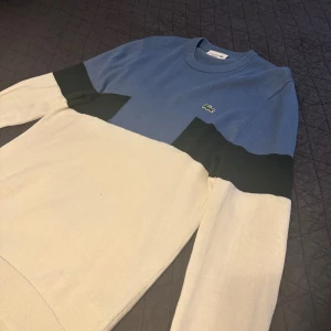 Blå och vit stickad tröja Lacoste - Snygg stickad tröja från Lacoste i blått och vitt med svarta detaljer över bröstet. Klassisk rund halsringning och långärmad modell. Ikoniska krokodilloggan på bröstet. Perfekt för dig som gillar stilren och sportig look.