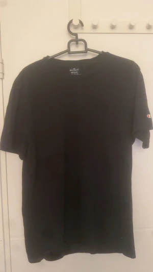 Svart t-shirt från Champion - En klassisk svart t-shirt från Champion i storlek M. T-shirten har rund halsringning, korta ärmar och den ikoniska Champion-loggan på ärmen. Tillverkad i mjuk bomull för en skön känsla hela dagen.