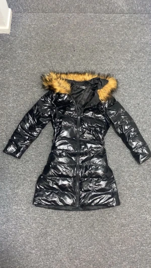 Svart lång pufferjacka med päls - Snygg svart lång pufferjacka med glansig finish och huva med fluffig brun fuskpäls. Jackan har quiltad design, dragkedja framtill och långa ärmar. Perfekt för kalla dagar när du vill hålla dig varm och samtidigt se cool ut.