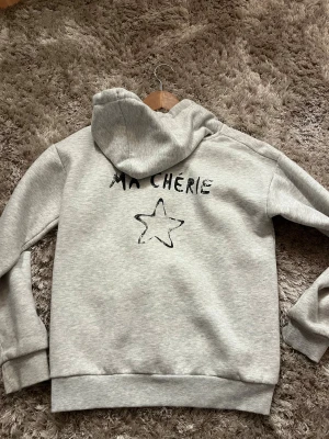 Grå hoodie med tryck 'Ma Chérie' och stjärna - Ljusgrå hoodie med huva och snörning. På bröstet och ryggen finns ett svart tryck med texten 'Ma Chérie' och en handritad stjärna. Tröjan har ribbade muddar vid ärmslut och nederkant. Perfekt för en chill och avslappnad stil.