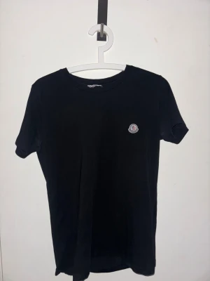 Svart Moncler t-shirt med logga - Svart t-shirt från Moncler med klassisk rund hals och korta ärmar. På vänster ärm sitter Monclers ikoniska broderade logga i vitt, rött och blått. T-shirten är gjord i mjuk bomull och har en clean, stilren look som funkar till allt. Det är storlek 1/S