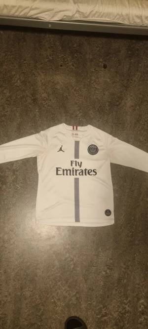PSG X JORDAN Vit långärmad - Säljer en vit långärmad PSG fotbollströja från Jordan med svart klubbmärke på bröstet och Fly Emirates-tryck. Tröjan har en diskret rand i mitten och är gjord i lätt, ventilerande funktionsmaterial. Perfekt för dig som älskar fotboll och vill sticka ut på planen. Passar mig perfekt som är (175cm-70kg)