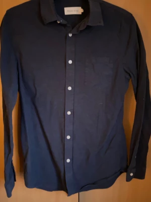 Mörkblå skjorta från Pier One - Snygg mörkblå skjorta från Pier One med klassisk krage och vita knappar framtill. Skjortan har en bröstficka och är långärmad, perfekt för en clean och stilren look. Materialet är mjuk bomull som känns skönt mot huden.