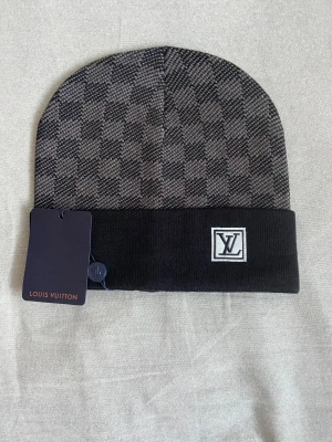 Louis Vuitton rutig svart mössa - Snygg mössa från Louis Vuitton med klassiskt rutigt mönster i svart och grått. Upptill är den stickad med diskret LV-logga på den svarta uppvikta kanten. Perfekt accessoar för dig som gillar exklusiv streetstyle och vill ha något som sticker ut.