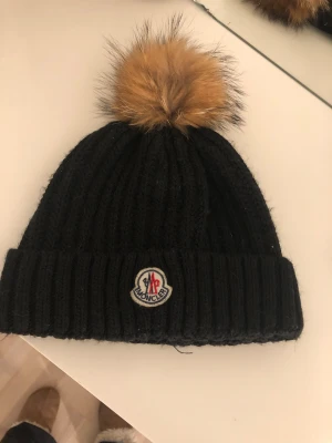 Svart Moncler mössa med pälsboll - Ribbstickad svart mössa från Moncler med stor brun pälsboll på toppen och klassisk Moncler-logga framtill. Perfekt för kalla dagar och ger en snygg vibe till vinterlooken. Mössan är mjuk och sitter skönt på huvudet.