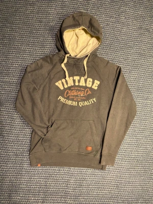 Blend Hoodie - Grå hoodie från Blend med beige insida på huvan och snören. Framsidan har ett stort vintage-inspirerat tryck i beige och brunt. Klassisk känguruficka framtill och ribbade muddar. Fet och bra basicplagg men med en bra vintagetuch. Om du undrar något är det bara att skriva🙌✅