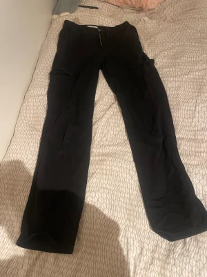 Svarta cargopants från STH - Svarta cargopants från STH med raka ben och stora sidofickor. Byxorna har dragkedja och knapp framtill, tillverkade i ett mjukt bomullsmaterial. Perfekt för dig som gillar streetwear och funktionella detaljer.