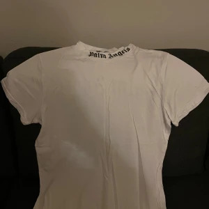 Vit Palm Angels t-shirt Street Wear S - Vit t-shirt från Palm Angles i storlek S med Palm Angels-tryck i svart på både kragen och ryggen. Klassisk rund hals och korta ärmar. T-shirten är gjord i mjuk bomull och har en clean streetstil som sticker ut med den feta loggan.
