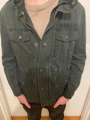 Grå/svart fieldjacka från Polo Ralph Lauren - Säljer en grön parkajacka från Polo Ralph Lauren i storlek 152. Jackan har fyra stora fickor med knappar, hög krage och coola detaljer som axelklaffar och USRL-tryck på bröstet. Materialet är slitstark bomull och passformen är normal.