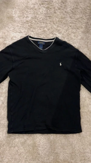 Svart långärmad tröja Polo Ralph Lauren - Svart långärmad tröja från Polo Ralph Lauren med vit broderad logga på bröstet och vit kant vid v-ringad hals. Tröjan är i mjuk bomull och har en klassisk, stilren look som funkar till jeans eller chinos. Perfekt för dig som gillar enkel och snygg design.