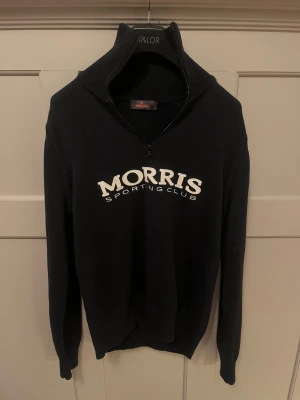 Svart stickad quarter zip från Morris - Svart stickad tröja från Morris Sporting Club med vit broderad logga på bröstet. Tröjan har hög krage med half zip-dragkedja och långa ärmar. Perfekt för dig som gillar klassisk och sportig stil.