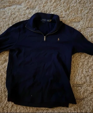 Morkblå quarter zip från Polo Ralph Lauren - Snygg marinblå långärmad tröja från Polo Ralph Lauren med klassisk broderad logga på bröstet. Tröjan har en stilren half zip-dragkedja och hög krage. Perfekt för lager-på-lager och passar till allt. Materialet är mjukt och skönt bomull.