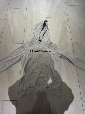 Grå hoodie från Champion. Pojke - Säljer en grå hoodie från Champion med svart logga på bröstet. Tröjan har huva, lång ärm och en stor magficka framtill. Perfekt för chill dagar eller när du vill ha en enkel och snygg streetstil.