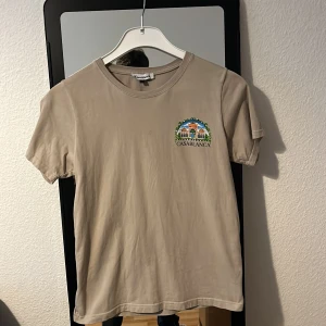 Beige t-shirt från Casablanca storlek 164 - Beige t-shirt från Casablanca med färgglada broderade och tryckta motiv fram och bak. Klassisk rund hals och korta ärmar. T-shirten är i mjuk bomull och har en relaxed vibe med snygg grafik och logga.