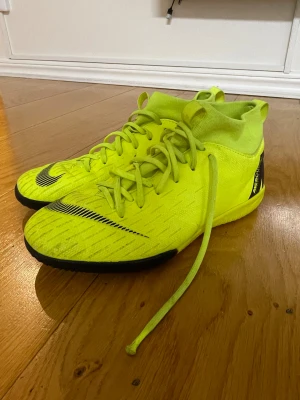 Neongula Nike Mercurial fotbollsskor - Säljer ett par neongula Nike Mercurial fotbollsskor med svart sula och snygga svarta detaljer. Skorna har snörning, platt sula och en strumpliknande krage för extra stöd. Perfekta för inomhusspel och har en modern, sportig look som sticker ut på planen.