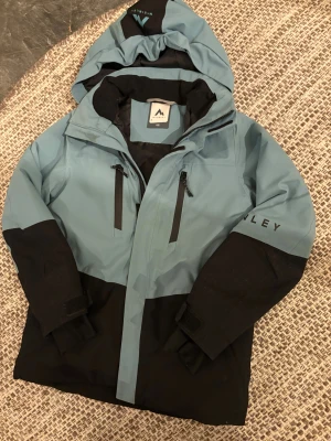 Blå och svart vindjacka från McKinley - Snygg vindjacka från McKinley i blått och svart med huva och två vertikala dragkedjor framtill. Jackan har justerbara muddar med kardborre och logga på ärmen. Perfekt för dig som gillar outdoor och vill ha en cool look.