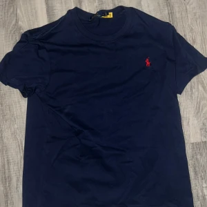 Mörkblå t-shirt från Ralph Lauren - Snygg mörkblå t-shirt från Ralph Lauren med klassisk röd logobrodering på bröstet. Modellen har rund hals och korta ärmar, perfekt för en clean och stilren look. Materialet är mjuk bomull som känns skönt mot huden.