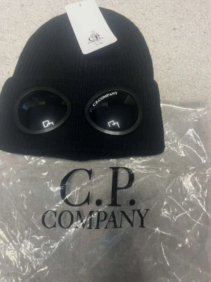Svart C.P. Company Goggle mössa - Svart ribbstickad mössa från C.P. Company med ikoniska goggle-detaljer framtill. Mössan har en uppvikt kant och är tillverkad i mjukt stickat material som ger en clean och modern look. Perfekt för dig som vill sticka ut med streetwear-vibe.