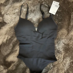 Svart body med tunna axelband - Snygg svart body med tunna, justerbara axelband och vadderade kupor. Djup v-ringning framtill och figurnära passform. Materialet är stretchigt och följsamt för en bekväm känsla.