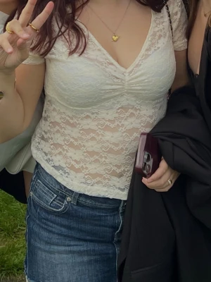 Vit spetsblus med v-ringning - Söt vit blus i spets med v-ringning och korta ärmar. Blusen har en lätt rynkning vid bysten och är genombruten med blommigt spetsmönster. Perfekt att styla med jeanskjol eller byxor.