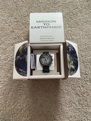 Omega x Swatch Mission to Earthphase - Snygg klocka från Omega x Swatch Bioceramic MoonSwatch Collection, modell Mission to Earthphase. Klockan har svart och grå urtavla med tachymeter och flera subdials. Svart armband med vit söm och Omega-logga. Levereras i originalförpackning.