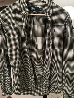Rutig skjorta från Polo Ralph Lauren - Klassisk svartvit rutig skjorta från Polo Ralph Lauren med button-down krage och broderad logga på bröstet. Skjortan har långa ärmar och knäppning framtill. Perfekt för en stilren och avslappnad look.