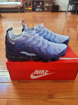 Nike Air Vapormax Plus blå/vit - Nike Air Vapormax Plus sneakers i en snygg gradient från vitt till blått. Ovandelen är i textil och syntet med vågiga detaljer, och sulan är svart med genomskinliga bubblor och vita stänk. Ikonisk Swoosh-logga på sidan och bekväm passform med snörning.