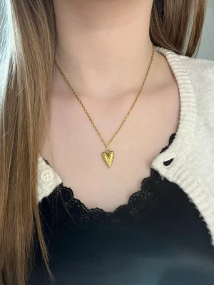 Guldigt halsband med hjärthänge - Snyggt halsband i guldfärg med ett hjärtformat hänge som har en pärlstruktur. Kedjan är tunn och elegant, perfekt för att ge en söt touch till din outfit. Hjärtat har en unik textur som gör smycket extra speciellt. Rostfritt stål och handgjort 