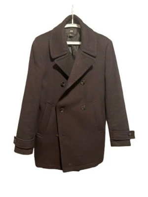 Svart skepparkavaj från H&M - Stilren svart skepparkavaj från H&M med dubbelknäppning, bred krage och två snedställda fickor framtill. Klassisk modell med knappar vid ärmslut och mjukt ullblandat material som håller värmen under kalla dagar.