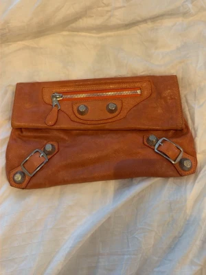 Korall Orange kuvertväska från Balenciaga - Säljer en så fin orange / korallfärgad kuvertväska från Balenciaga i läder. Väskan är vintage och har silverfärgade nitar, dragkedja framtill och dekorativa spännen. Perfekt nu till våren/sommaren, skulle även vara perfekt för balen.