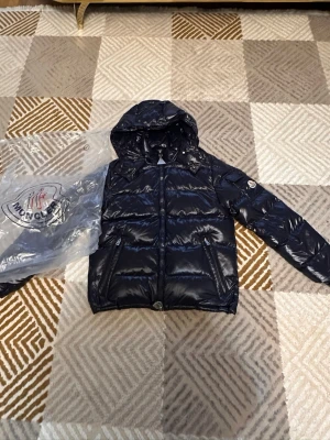 NFC fungerar Svart dunjacka med huva från Moncler - Snygg svart dunjacka från Moncler med glansig finish och huva. Jackan har dragkedja framtill, två fickor med dragkedja och Moncler-logga på ärmen. Perfekt för kalla vinterdagar och riktigt trendig stil. 