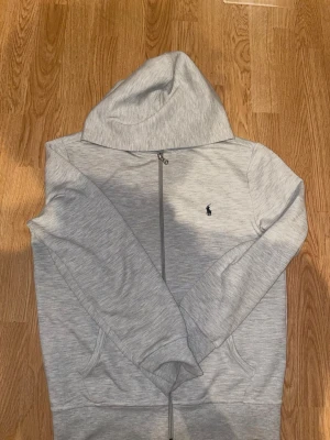 Ljusgrå hoodie från Polo Ralph Lauren - Snygg ljusgrå hoodie med dragkedja från Polo Ralph Lauren. Klassisk design med huva, två fickor framtill och den ikoniska lilla loggan broderad på bröstet. Perfekt för en avslappnad och stilren look. Utan snören!!!
