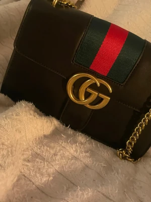 Gucci svart axelremsväska med kedja - Snygg svart handväska kopia Gucci i läder med klassiska gröna och röda ränder på locket. Väskan har en guldig kedjerem och GG-logga framtill. Perfekt för dig som vill ha en ikonisk och trendig accessoar med lyxig känsla.