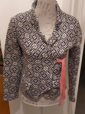 Mönstrad stickad kofta med rosa band - Snygg stickad kofta, från Oddmolly,med svartvitt geometriskt mönster och rosa knytband i midjan. Koftan har lång ärm, hög krage och dekorativa rosa knappar bak. 