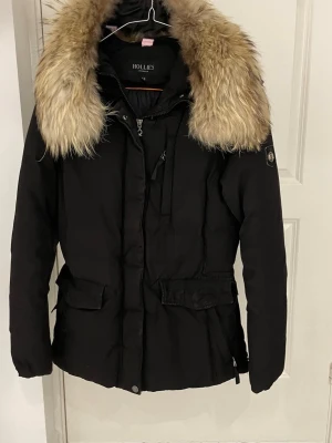 Svart dunjacka från Holliester med äktapäls  - Svart dunjacka från Holliester med stor, fluffig äktapäls i beige runt kragen. Jackan har dragkedja och knappar framtill, två stora fickor med lock och en figurnära passform. Perfekt för kalla dagar och riktigt snygg streetstil. Bra skick men har tappat lite färg.
