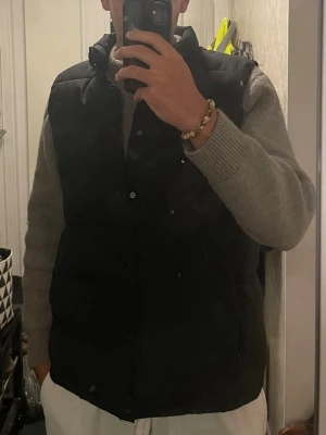 Svart dunväst - Snygg svart dunväst med ståkrage och knäppning framtill. Västen har en klassisk, puffig design och är perfekt att slänga över en hoodie eller tröja för extra värme. I storlek L från construe 