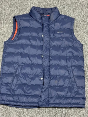 Marinblå dunväst från GANT - Snygg marinblå dunväst från GANT med orange foder och diskret logga på bröstet. Västen har ståkrage, dragkedja och tryckknappar framtill samt två snedställda fickor. Perfekt för lager-på-lager och ger en sportig look.