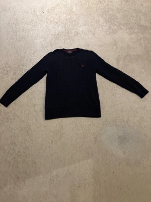 Mörkblå kabelstickad tröja Polo Ralph Lauren - Mörkblå kabelstickad tröja från Polo Ralph Lauren med rund halsringning och röd broderad logga på bröstet. Klassisk modell med ribbstickade muddar och nederkant. Perfekt för en stilren look.