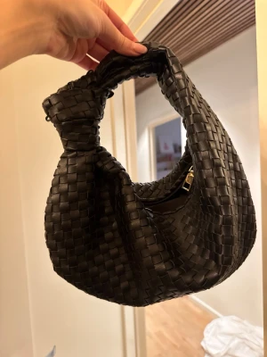 Svart flätad axelväska i läderimitation - Trendig svart handväska med flätad design och rundad form. Väskan har en kort axelrem med knutdetalj och dragkedja i guld. Tillverkad i läderimitation som ger en lyxig känsla. Perfekt för dig som gillar stilrena accessoarer.