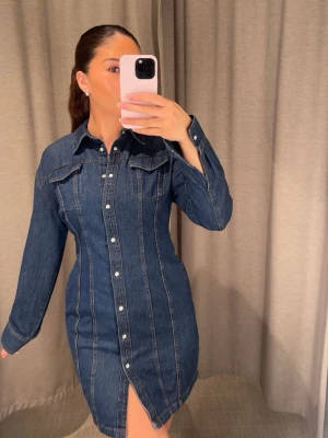 Jeansklänning  - Snygg jeansklänning i mörkblå denim med långa ärmar, knappar framtill och fickor. Använd en gång. Säljer pågrund av att jag har en liknande jenasklänning. 